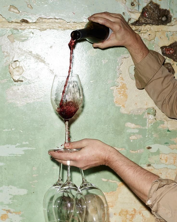 A hand pouring red wine into a glass against a weathered green wall